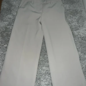 Beige vida kostymbyxor - Stilrena beige kostymbyxor med vida ben och klassisk passform. Byxorna har hällor för bälte och pressveck framtill, vilket ger en clean och dressad look. Perfekta för dig som gillar en enkel men trendig stil.