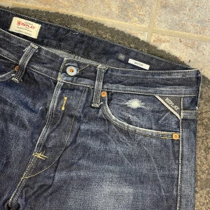 Replay Waitom jeans - Snygga Replay Waitom jeans i mörkblå tvätt med slitningar och ljusa detaljer. Klassisk femficksmodell med kontrastsömmar och Replay-logga på fickan. Jeansen har rak passform och är tillverkade i robust denim för en cool vardagslook.