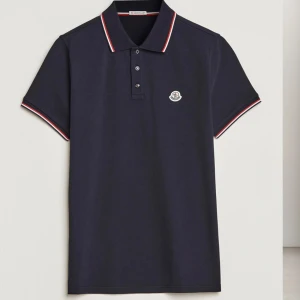 Mörkblå Moncler pikétröja - Snygg mörkblå pikétröja från Moncler med klassisk krage och knappar framtill. Tröjan har röda och vita detaljer på krage och ärmslut samt Moncler-logga på bröstet. Perfekt för en stilren och sportig look.