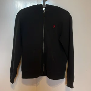 Svart hoodie från Polo Ralph Lauren - Svart hoodie från Polo Ralph Lauren med dragkedja framtill och klassisk röd logga broderad på bröstet. Inga defekter, otroligt bra skick.Tröjan har huva, fickor och långa ärmar. Perfekt för en chill och stilren look. Kom privat vid minsta fundering! Passar även S. Kan gå ner i pris vid snabb affär!