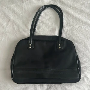 Svart handväska - Stilren svart axelväska med rundade kanter och dubbla handtag. Väskan har detaljer i skinnimitation och stängs med dragkedja upptill. Insidan har flera fack på insidan, inklusive ett med dragkedja, perfekt för att organisera dina grejer. Aldrig använd, nyskick!