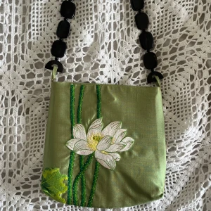 Grön axelväska med blommigt broderi - Unik grön axelväska i siden med broderad vit blomma och gröna stjälkar på framsidan. Väskan har en cool axelrem i svarta plastpärlor Perfekt för dig som vill ha en clutch, eller bara en gullig detalj till din outfit då utrymmet är rätt litet och platt. 