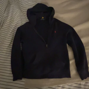 Mörkblå hoodie från Polo Ralph Lauren - Snygg mörkblå hoodie från Polo Ralph Lauren med dragkedja och klassisk röd logga på bröstet. Tröjan har huva, långa ärmar och är tillverkad i mjuk bomull. Perfekt för en chill och stilren look.