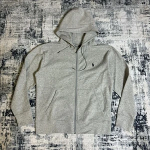 Grå zip Hoodie från Polo Ralph Lauren - Nypris 1900kr. Snygg grå hoodie från Polo Ralph Lauren med dragkedja framtill och klassisk huva med snörning. Tröjan har två fickor och den ikoniska lilla broderade loggan på bröstet.