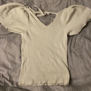 Vit ribbad topp med puffärm från ONLY - Supersöt vit ribbad topp från ONLY med v-ringning, korta puffärmar och knytning i nacken. Toppen har en figurnära passform och är perfekt för dig som gillar en feminin och trendig stil. Materialet är mjukt och stretchigt för extra komfort.