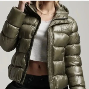 Olivgrön pufferjacka med huva - Säljer en olivgrön pufferjacka med glansig finish och huva. Jackan har breda, quiltade kanaler, dragkedja framtill och två fickor. Perfekt för kalla dagar och ger en cool streetstyle-vibe. Passformen är normal och jackan är långärmad.priset går att diskutera