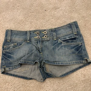 Jeansshorts från Kate Moss - Snygga blå jeansshorts från Kate Moss med dubbla knappar framtill och klassiska fickor både fram och bak. Shortsen har låg midja och coola slitningar för en avslappnad look. Perfekta för varma dagar och festivaler. Midjemått 38 cm