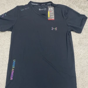 Svart Under Armour HeatGear t-shirt - Svart t-shirt från Under Armour med färgglatt logotryck på bröstet och texten UNDER ARMOUR i regnbågsfärger längs sidan. Gjord i lätt och teknisk polyester med HeatGear-teknologi som håller dig sval. Perfekt för löpning eller träning.