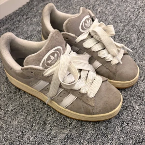 Adidas Campus grå mocka sneakers - Grå Adidas Campus sneakers i mocka med vita detaljer och klassiska tre ränder på sidorna. Skorna har vit sula med gummidetalj, rund tå och breda vita snören. Adidas logga på plös och häl. Perfekta för en clean och stilren look.