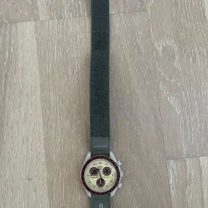 Omega x Swatch MoonSwatch Pluto - Säljer en Omega x Swatch MoonSwatch Mission to Pluto. Klockan har en beige urtavla med vinröda detaljer och grått band med Omega- och Swatch-logga. Boetten är vit och klockan har kronograf-funktion. Perfekt för dig som gillar snygga och unika accessoarer.
