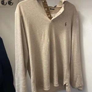 Beige pikétröja från Polo Ralph Lauren - Snygg beige långärmad pikétröja från Polo Ralph Lauren i custom slim fit. Tröjan har klassisk krage, knappar framtill med kontrast i brunt och den ikoniska broderade loggan på bröstet. Perfekt för en clean och stilren look.