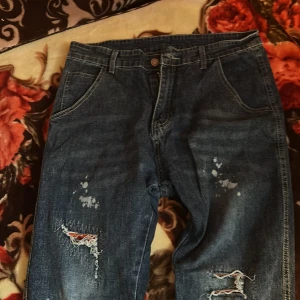 Blå jeans med slitningar och stänk - Säljer ett par blå jeans med coola slitningar och färgstänk på benen. Jeansen har raka ben, klassiska fickor och en patch på bakfickan. Perfekt för dig som gillar en avslappnad och trendig streetstil.
