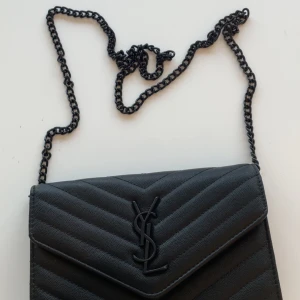 Svart quiltad axelväska från Saint Laurent - Säljer en svart quiltad axelväska från Saint Laurent med ikoniskt YSL-emblem framtill. Väskan har kedjerem i svart metall, flera fack inuti med röd insida och dragkedja. Tillverkad i skinn med elegant struktur och klassisk kuvertform.