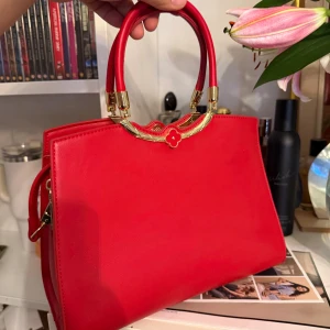 Röd handväska med gulddetaljer - Snygg röd handväska i skinn med stilrena gulddetaljer och dekorativt handtag. Väskan har en klassisk, fyrkantig form och ett elegant blommotiv i metall vid handtaget. Perfekt accessoar för att lyfta din outfit.