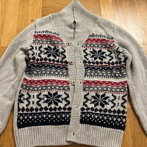 Grå kofta med mönster från Hampton Republic - Snygg grå stickad kofta från Hampton Republic med klassiskt vintermönster i svart, vitt och rött framtill. Koftan har ribbade muddar, knappar och hög krage. Perfekt för kalla dagar när du vill ha en mysig och stilren look.