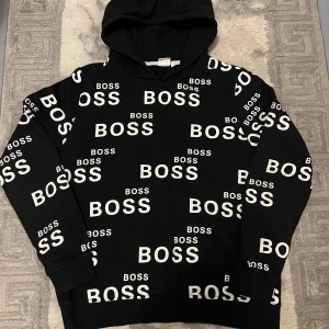Svart hoodie från Hugo Boss med print - Svart hoodie från Hugo Boss gammal model  i storlek M 16 år  med vit BOSS-logga tryckt över hela tröjan. Tröjan har huva och långa ärmar, tillverkad i mjuk bomull. Perfekt för dig som gillar streetstyle och vill sticka ut med ett tydligt märke.