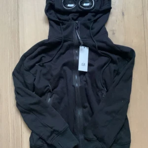 Svart hoodie med glasögon  - Svart hoodie med dragkedja och ikoniska glasögon i huvan. Tröjan har långa ärmar, ribbade muddar och två fickor med dragkedja. Perfekt för dig som vill sticka ut med en unik streetwear-look.