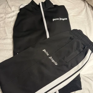 Palm Angels Tracksuit  - Svart från Palm Angels Tracksuit med vita sidoränder och logga tryckt på låret och bröstet . Byxorna har elastisk midja och raka ben, tillverkade i ett mjukt och sportigt material. Perfekta för en chill och trendig streetwear-look.