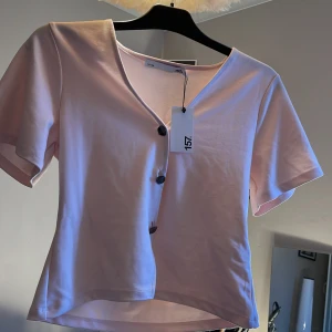 Ljusrosa blus med knappar från 157 - Aldrig andvänd.  Supersöt ljusrosa blus från 157 med korta ärmar och v-ringning. Blusen har en unik knappslå med svarta knappar som går snett framtill. Tillverkad i mjuk bomull, perfekt för en fräsch och enkel look.