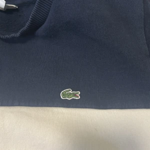 Färgglad Lacoste sweatshirt i blockfärger - Säljer en snygg Lacoste sweatshirt med blockfärger i marinblått, vitt och rött. Tröjan har klassisk rund hals, lång ärm och broderad krokodil-logga på bröstet samt Lacoste-tryck framtill. Perfekt för dig som gillar streetstyle och vill sticka ut.