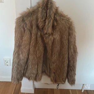 Brun pälsjacka med fluffig look - Säljer en brun pälsjacka med lång, fluffig fuskpäls och hög krage. Jackan har en klassisk rak passform och är perfekt för dig som vill sticka ut med en trendig och mysig stil. Fodrad insida för extra komfort.