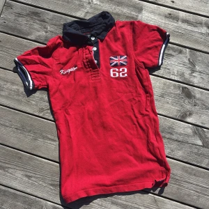 Röd Kingston t-shirt med krage XXS - Snygg röd t-shirt från Kingston med svart krage och vita knappar. Framsidan har broderad Union Jack-flagga och siffran 62, samt Kingston-logga. Ärmarna är korta med vit och svart kant. Baksidan har en vit och svart rand längs ryggen. Materialet är bomull.