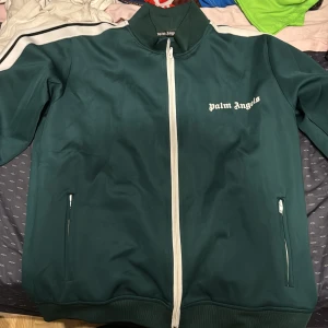 palm angels grön zip - Säljer en grön track jacket från Palm Angels med vita detaljer och dragkedja framtill. Jackan har ståkrage, vita ränder längs ärmarna och logga på bröstet. Perfekt för en sportig och trendig look.