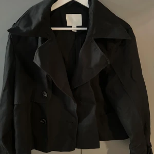 Svart kort trenchcoat - Svart kort trenchcoat med breda slag och dubbelknäppning framtill. Den slutar typ vid midjan beroende på hur lång man är (jag är 170 cm)