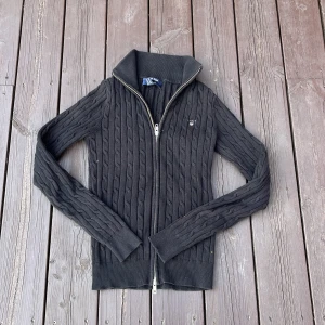 Svart kabelstickad kofta från Gant - Svart kabelstickad kofta från Gant i stretchig bomull. Tröjan har hel dragkedja framtill, hög krage och ribbade muddar. Klassisk och stilren design med Gant-logga på bröstet. Perfekt för lager-på-lager och snygg till jeans eller kjol.