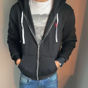 Svart hoodie från Polo Ralph Lauren - Svart hoodie från Polo Ralph Lauren med vit snörning och dragkedja framtill. Slim fit-modell med klassisk huva och fickor på magen. Liten röd logga broderad på bröstet. Perfekt för en chill och stilren look.