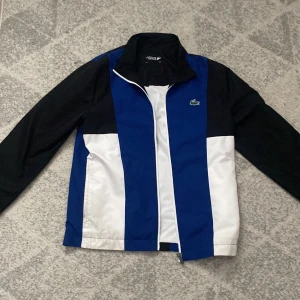 Blå och vit vindjacka från Lacoste Sport - Snygg vindjacka från Lacoste Sport i blått, vitt och svart med klassisk krokodillogga på bröstet. Jackan har hel dragkedja framtill, ståkrage och långa ärmar. Perfekt för dig som gillar sportig och clean stil. Priset kan diskuteras 