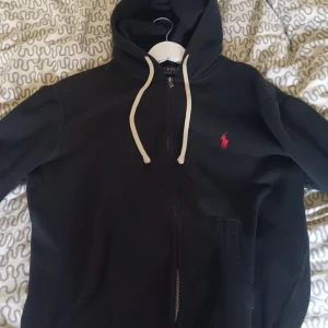 Svart hoodie från Polo Ralph Lauren - Svart hoodie från Polo Ralph Lauren med dragkedja och vit snörning i huvan. Klassisk röd broderad logga på bröstet och känguruficka framtill. Perfekt för en chill och stilren look.