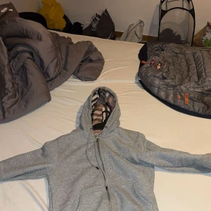 Grå zip-hoodie från Burberry - Säljer en grå zip-hoodie från Burberry med klassiskt rutigt foder i huvan och insidan. Jackan har dragkedja, snörning i huvan och broderad Burberry-logga på bröstet. Perfekt för dig som vill ha en stilren och lyxig hoodie med ikoniska detaljer.