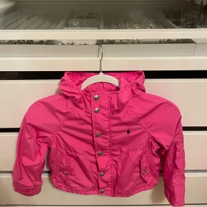 Rosa vindjacka från Ralph Lauren - Snygg rosa vindjacka från Ralph Lauren med huva, dragkedja, tryckknappar framtill, två fickor med knappstängning och klassisk Polo-logga broderad på bröstet. Nypris över 1300kr!