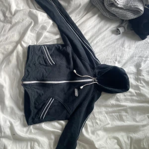 Svart hoodie med dragkedja från DKNY - Svart hoodie från DKNY i storlek M med vit dragkedja och vita detaljer längs fickor och ärmar. Tröjan har huva med snörning och två fickor framtill. Perfekt för en chill och sportig look.