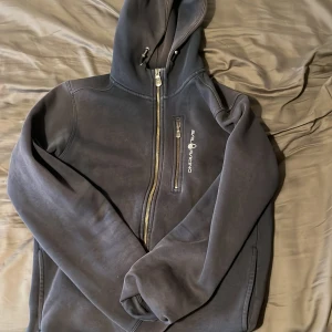 Mörkblå hoodie från Sail Racing - Snygg mörkblå hoodie från Sail Racing med dragkedja och huva. Tröjan har en bröstficka med dragkedja och Sail Racing-logga på bröstet. Perfekt för kyliga dagar och har en skön, mjuk insida. Klassisk passform och stilren look.