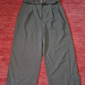 Grå croppade wide byxor från H&M - Snygga grå byxor från H&M i croppad modell med breda ben och hög midja. Byxorna har bälte i midjan, sidofickor och bakfickor med knapp. Perfekta för dig som gillar en chill och trendig stil. Materialet är mjukt och känns skönt mot huden.Khaki färg