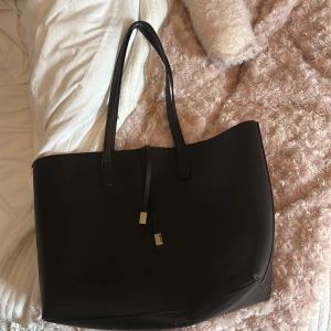 Svart shoppingväska i skinn - Stilren svart shoppingväska med långa handtag och minimalistiska metalldetaljer. Väskan är rymlig och har en enkel, modern design som passar perfekt för att bära allt du behöver under dagen.