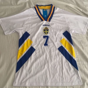 Svensk landslagströja Larsson #7 Adidas M - Säljer en klassisk svensk landslagströja från Adidas i storlek M, med Larsson och nummer 7 på ryggen. Tröjan är vit med blå och gula detaljer, V-ringad krage med blå, gul och röd kant. Tillverkad i 100% polyester, perfekt för fotbollsfans. Den är aldrig använd. Nypris 1599 kr.