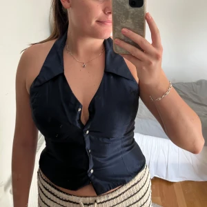 Mörkblå halterneck korsett med knappar - Snygg mörkblå korsett-topp med halterneck och stora slag vid kragen. Toppen har knäppning framtill med glansiga knappar och är figurnära med öppen rygg. Perfekt för dig som vill ha en trendig och stilren look.