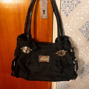 Svart handväska från Gamaza med detaljer - Snygg svart handväska från Gamaya med glansiga handtag och coola silvriga metalldetaljer. Väskan har ett diskret mönster med G-loggor och blommor i tyget samt en metallplatta med strass framtill. Perfekt för dig som gillar statement-accessoarer.