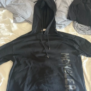 Svart hoodie från Moncler med tryck - Svart hoodie från Moncler med stort tryck längs sidan framtill. Klassisk huva med snörning och relaxed fit. Tillverkad i mjuk bomull, perfekt för chill dagar eller när du vill ha en clean streetstyle-look. Pris kan diskuteras 