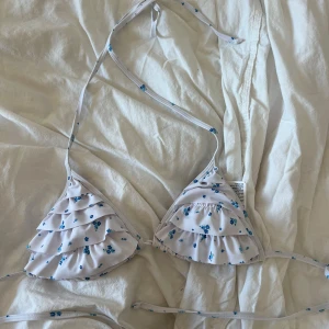 Vit blommig volangbikini från Good Collection - Supersöt trekantsbikini i vitt med små blå blommor och volangdetaljer på kuporna. Knytband i nacken och ryggen , aldrig använd 