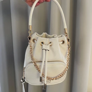 Vit bucket bag från ALDO med kedjedetalj - Säljer en snygg vit bucket bag från ALDO med guldfärgade metalldetaljer och kedja. Väskan har snörning upptill, ett handtag och en justerbar axelrem. Framtill finns en praktisk ficka med dragkedja. Perfekt för dig som vill ha en trendig och stilren väska. Köpt från Zalando, nypris 729kr. Mycket sparsamt använd.