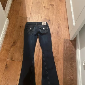 Mörkblå bootcut jeans från True Religion - Säljer ett par mörkblå jeans från True Religion med bootcut passform. Jeansen har snygga kontrastsömmar, ikoniska fickdetaljer med lock och silverknappar bak samt låg midja. Färgen är en aning urtvättad men fortfarande i bra skick annars!