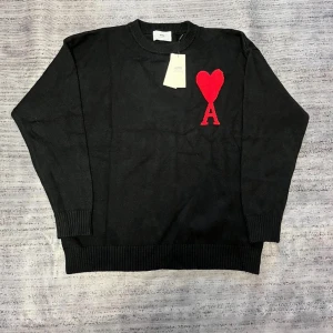 Svart stickad tröja från Ami Paris - Svart stickad tröja från Ami Paris med rund hals och ribbade muddar. På bröstet finns ett stort rött hjärta med bokstaven A. Perfekt för dig som gillar stilrena plagg med en twist. Materialet är mjuk bomull som känns skönt mot huden.