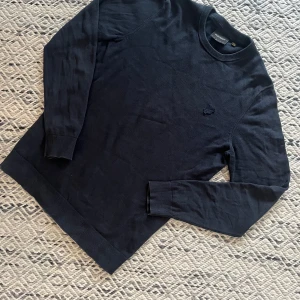Mörkblå stickad tröja från Lyle & Scott - Mörkblå stickad tröja från Lyle & Scott med rund halsringning och diskret broderad logga på bröstet. Tröjan har långa ärmar och ribbade muddar vid ärmslut och nederkant. Perfekt för dig som gillar en clean och stilren look.