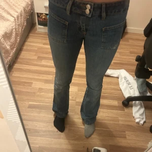 Blå bootcut jeans från Diesel - Säljer ett par snygga blå jeans från Diesel med bootcut passform och coola stora fickor fram. Jeansen har dubbla knappar i midjan, klassisk Diesel-logga och detaljerade sömmar. Perfekt för dig som gillar en avslappnad men trendig stil.