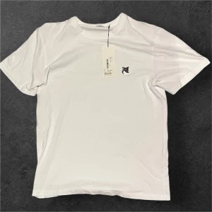 Maison Kitsuné vit t-shirt med tagg kvar - Taggen fins kvar, nypris 999kr på nk gbg aldrig använd! Vit t-shirt från Maison Kitsuné i bomull, med klassisk rund hals och korta ärmar. På bröstet sitter en broderad patch med ett grått rävhuvud som ger en clean men unik vibe. Perfekt för dig som gillar stilrena plagg med en twist.