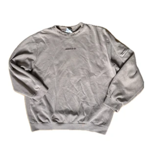 Beige sweatshirt från Adidas - Snygg beige sweatshirt från Adidas med broderad logga på bröstet och tryckt Adidas-detalj på kragen. Tröjan har långa ärmar, ribbade muddar och en patch på ena ärmen. Perfekt för en chill och sportig look.